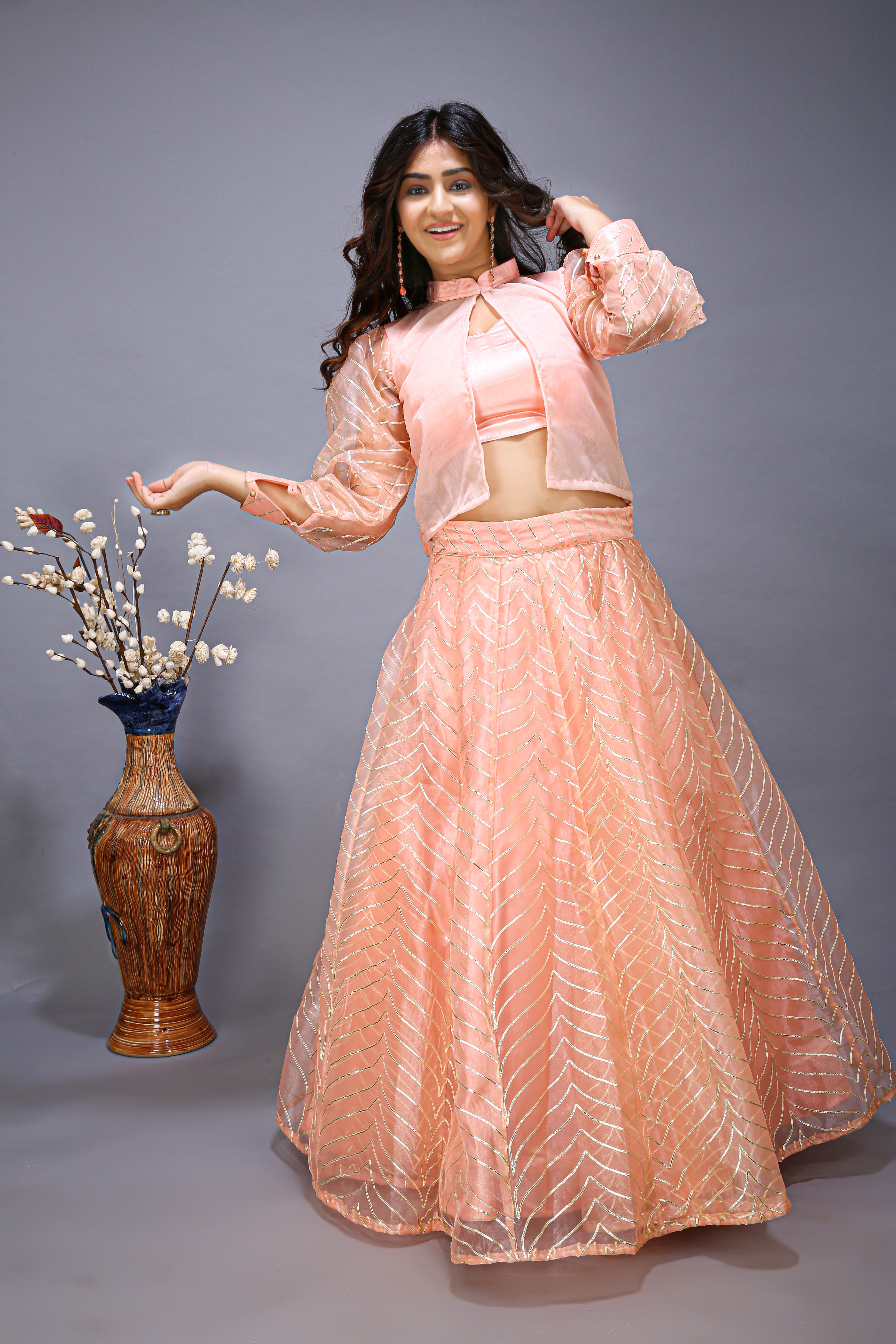 A woman posing in a pink lehenga choli with a stylish shrug, standing next to a vase with flowers.